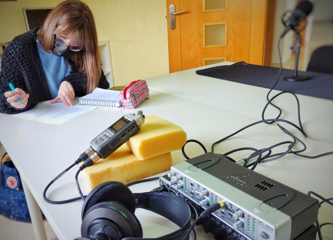 Momento previo á gravación do podcast. Sara Fraga repasa os seus apuntamentos, na mesa na que está o material de gravación, ordenado por Jorge Lama e listo para empregar: micrófono, auriculares e distribuidor de audio.